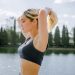 A woman adjusts her hair, getting ready for an outdoor workout by a lake on a sunny day.